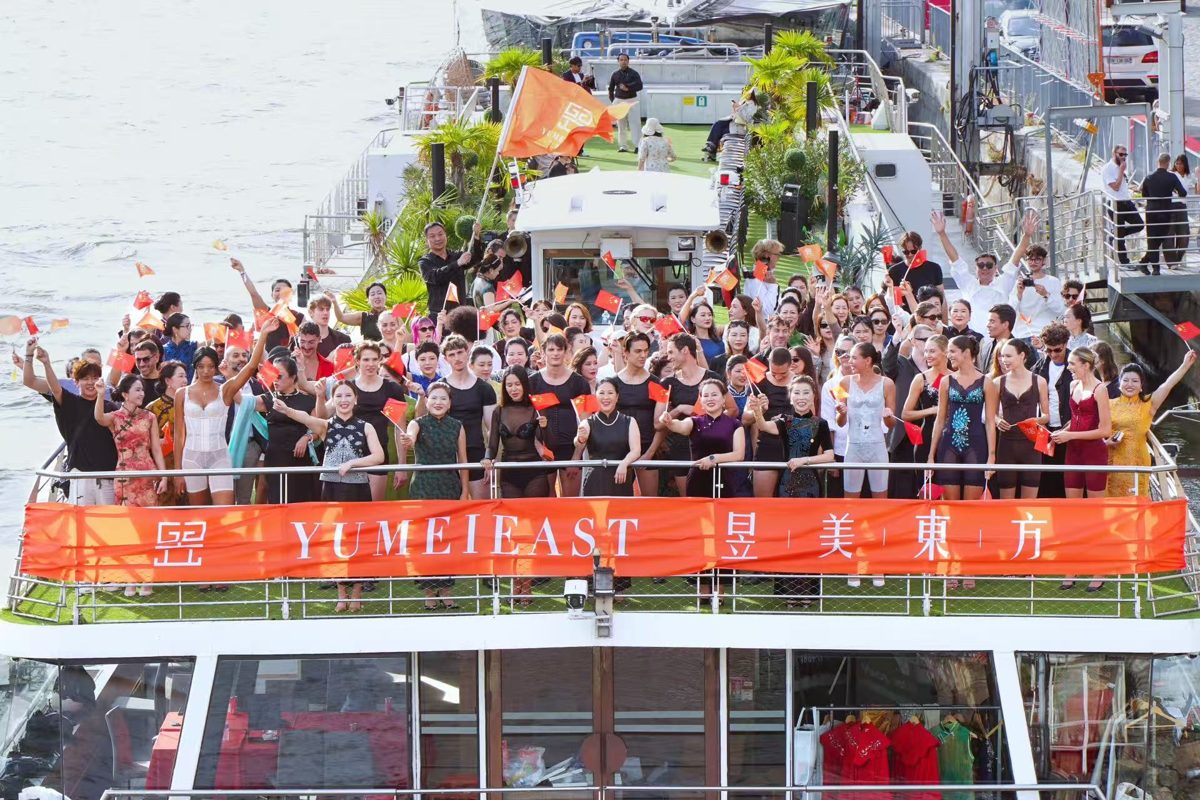 Guests and models waving flags on the deck of a boat decorated with a YuMei East banner on the Seine